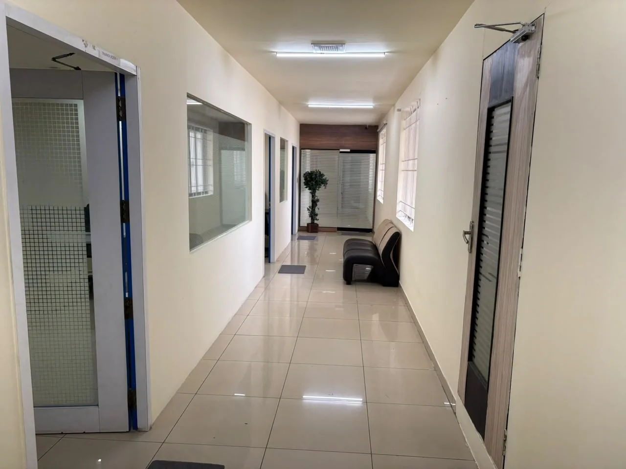 A long, brightly lit corridor with a tiled floor, glass-panelled doors on the left, windows on the right and a black waiting bench against the wall.