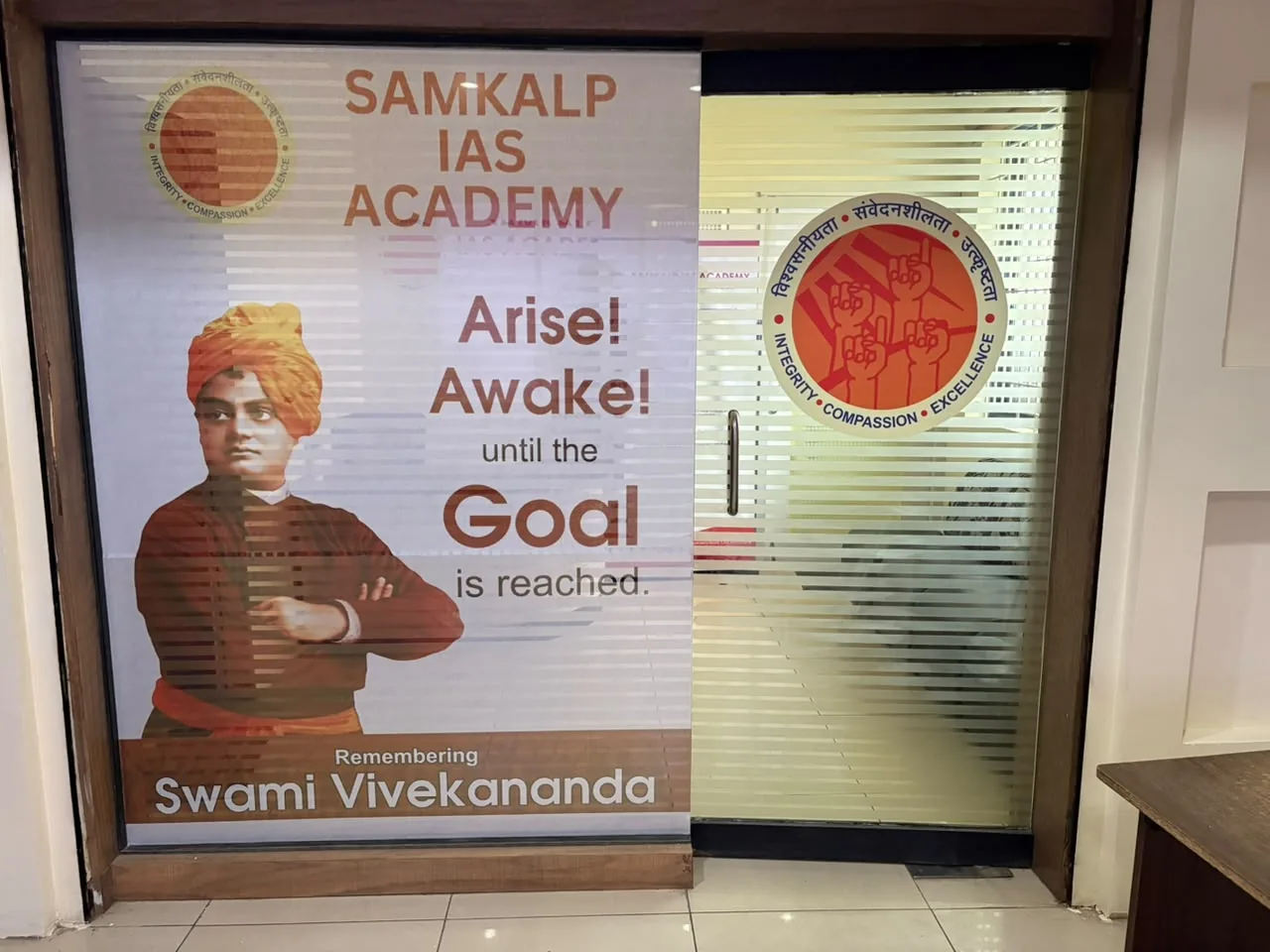 Glass door with horizontal frosted stripes. On the left panel a poster of Swami Vivekananda with the words Arise! Awake! until the Goal is reached; on the door the round orange academy emblem.