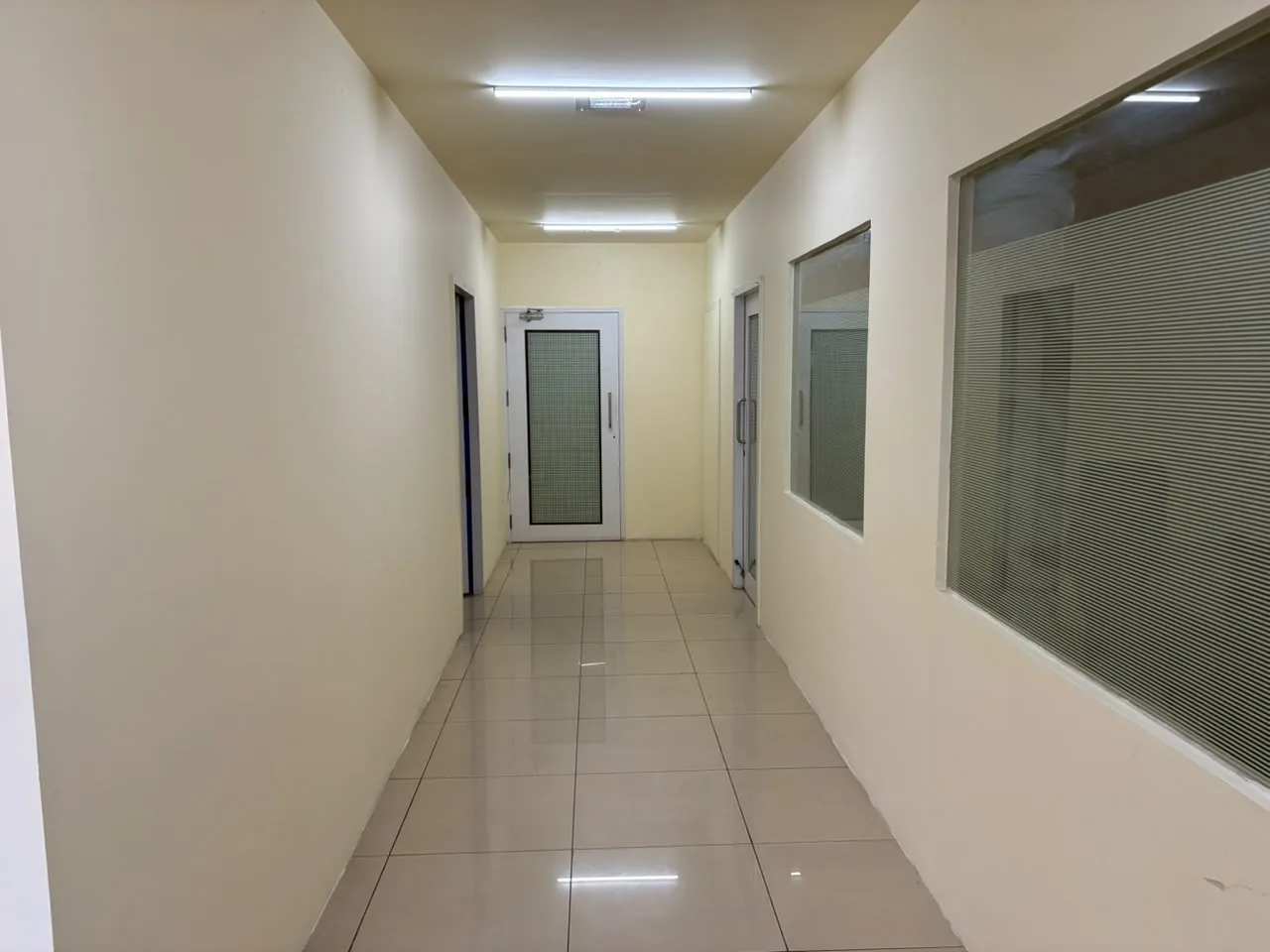 A narrow corridor with cream walls and a tiled floor ending at a white door with a frosted glass panel; a frosted window on the right.