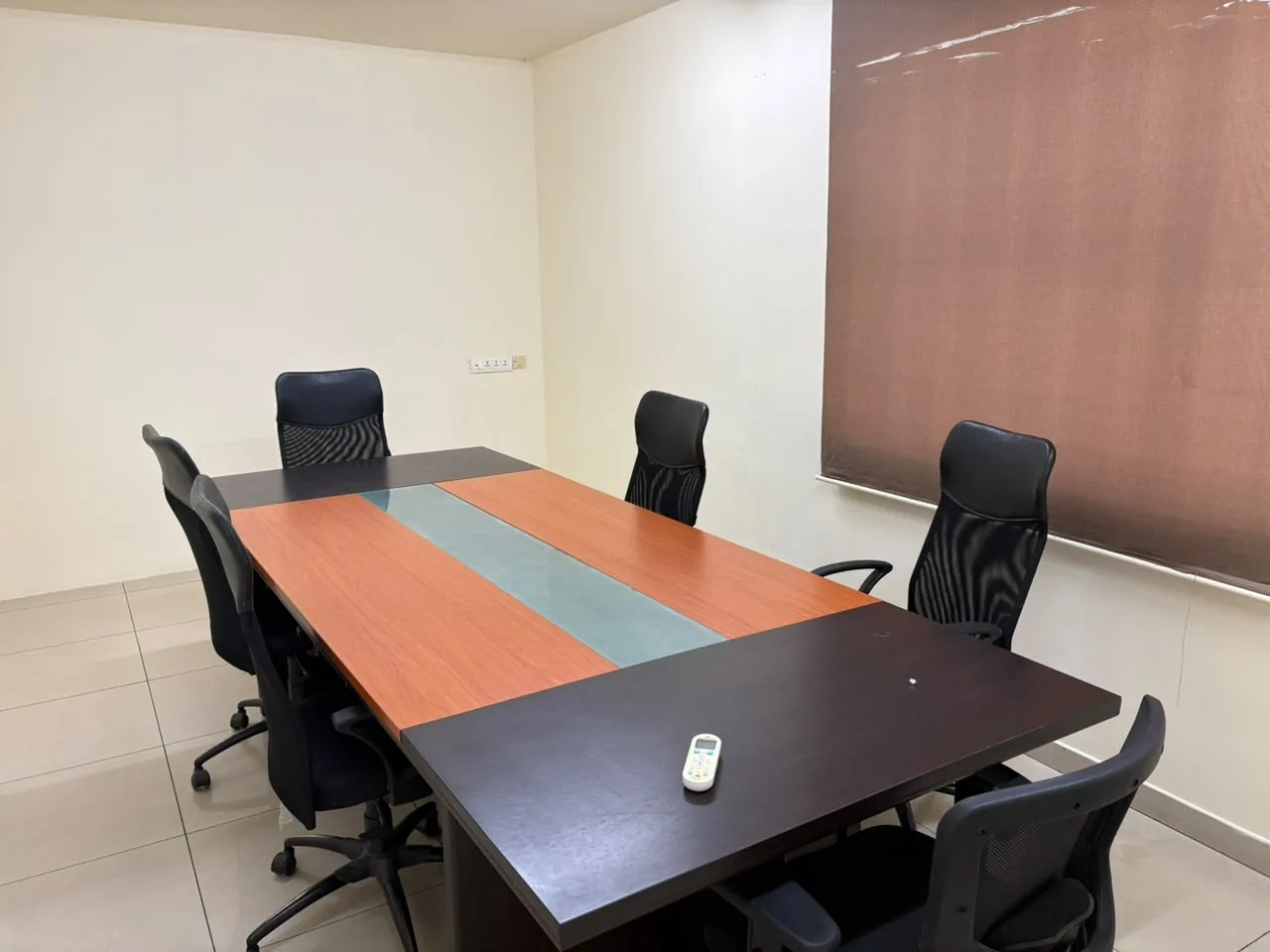 A meeting room with a rectangular wooden table, six black mesh office chairs and a brown window blind.