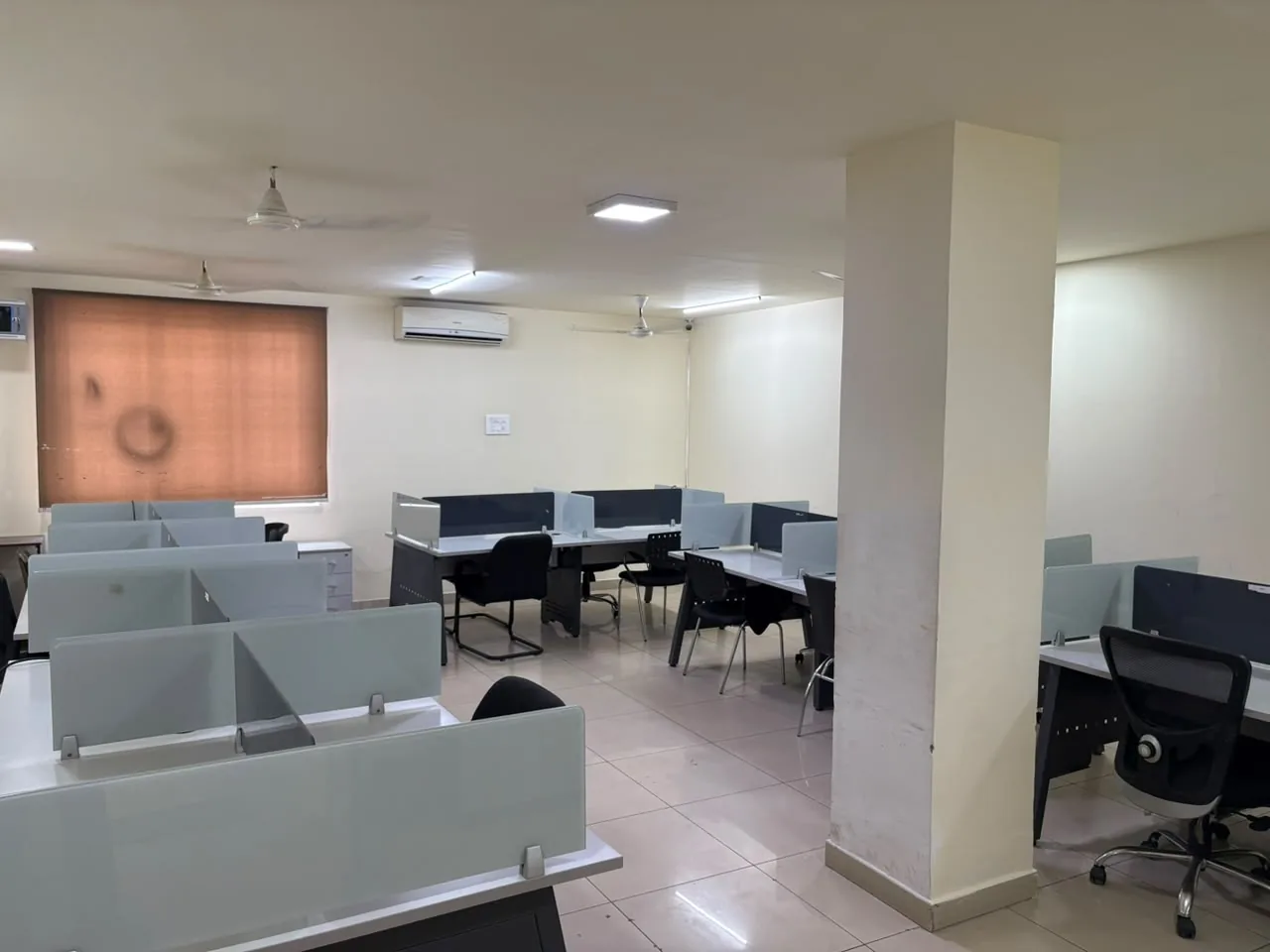 A large room with rows of white desks separated by glass partitions, office chairs, ceiling fans, a wall air conditioner and an orange window blind.