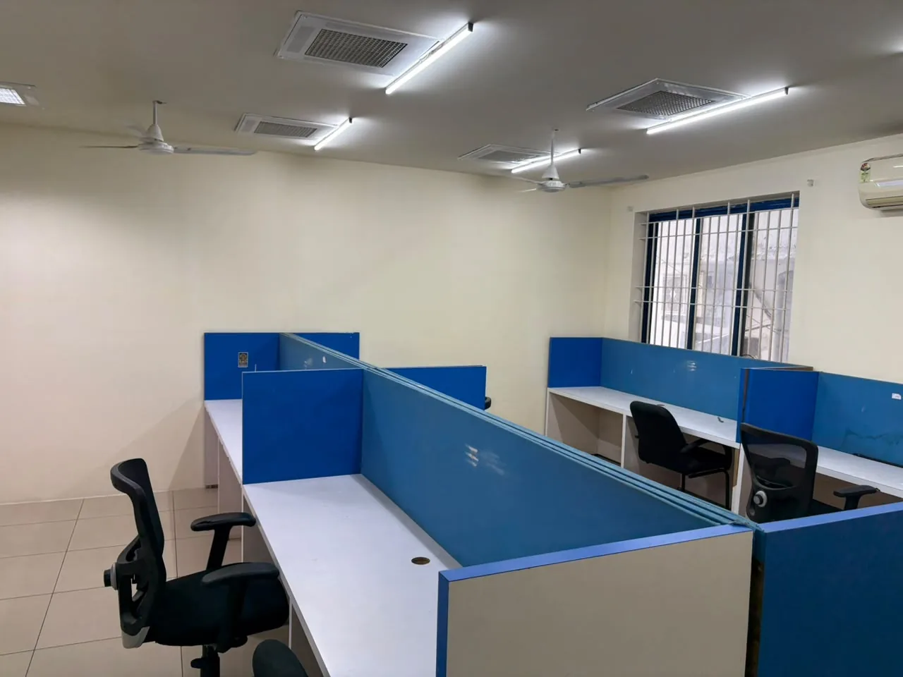 A room with white desks divided by blue partitions, black office chairs, ceiling fans, air-conditioning vents and a barred window.