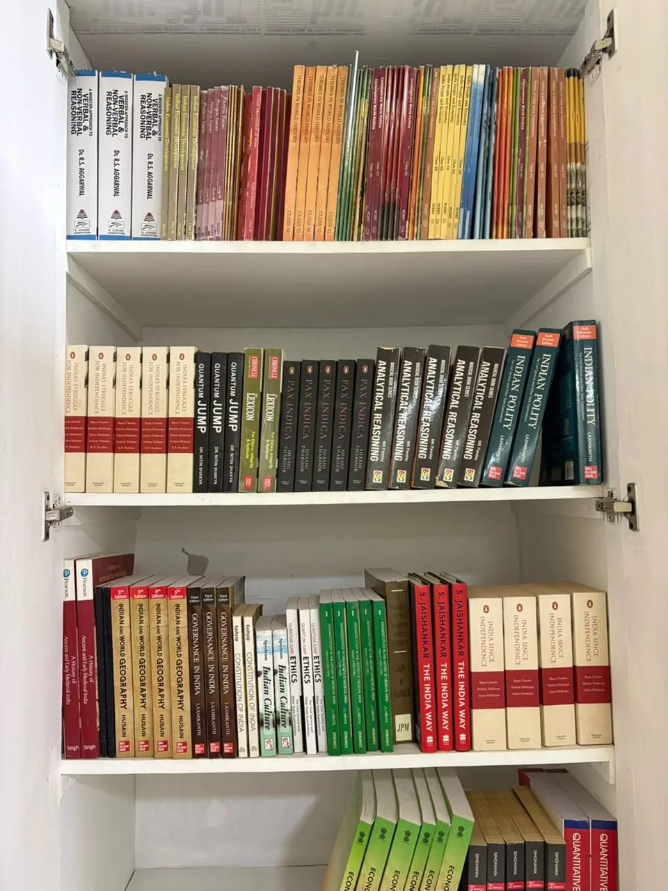 A white bookshelf with NCERT textbooks and reference books including Indian Polity by Laxmikanth, India's Struggle for Independence, India Since Independence and Indian and World Geography.